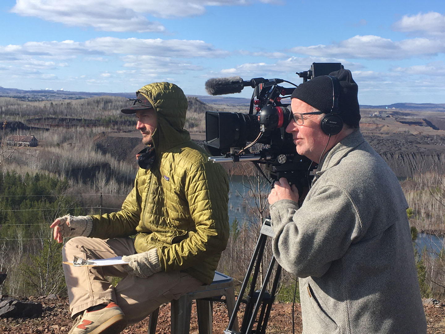 A camera man and reporter outside wearing warm clothes with trees and a horizon in the background